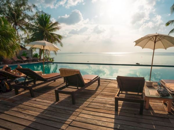 A serene poolside scene with lounge chairs, umbrellas, and a beautiful view of the ocean under a partially cloudy sky with sunlight breaking through.