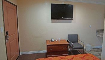 The image shows a modest hotel room with a TV mounted on the wall, a small table, a chair, and part of a bed visible at the bottom.