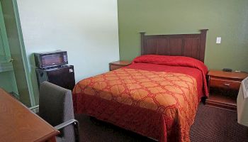 The image shows a small hotel room with a bed, bedside tables, a desk, chair, mini-fridge, and microwave. The walls are green, and the bedspread is red.