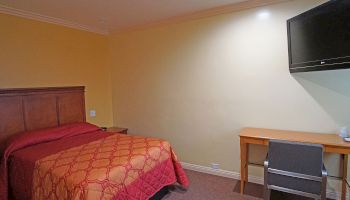 A bedroom with a single bed covered in a red quilt, a wooden desk, a chair, and a wall-mounted TV is shown in this image.