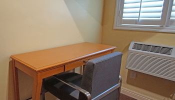 The image shows a small room corner with a wooden desk, a cushioned chair, a window with blinds, and a wall-mounted air conditioning unit.