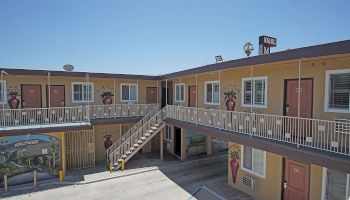 The image depicts a two-story motel with exterior corridors, orange doors, and murals of vases with flowers on the walls, under a clear blue sky.