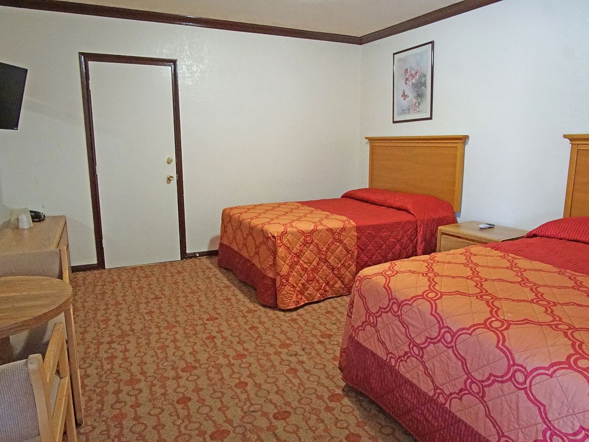 The image shows a hotel room with two double beds, a wall-mounted TV, a small wooden table with chairs, a dresser, and framed artwork on the wall.