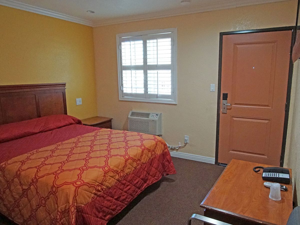 A small hotel room with a double bed, an orange bedspread, a window, and a tan door. A desk holds a phone and TV remote.