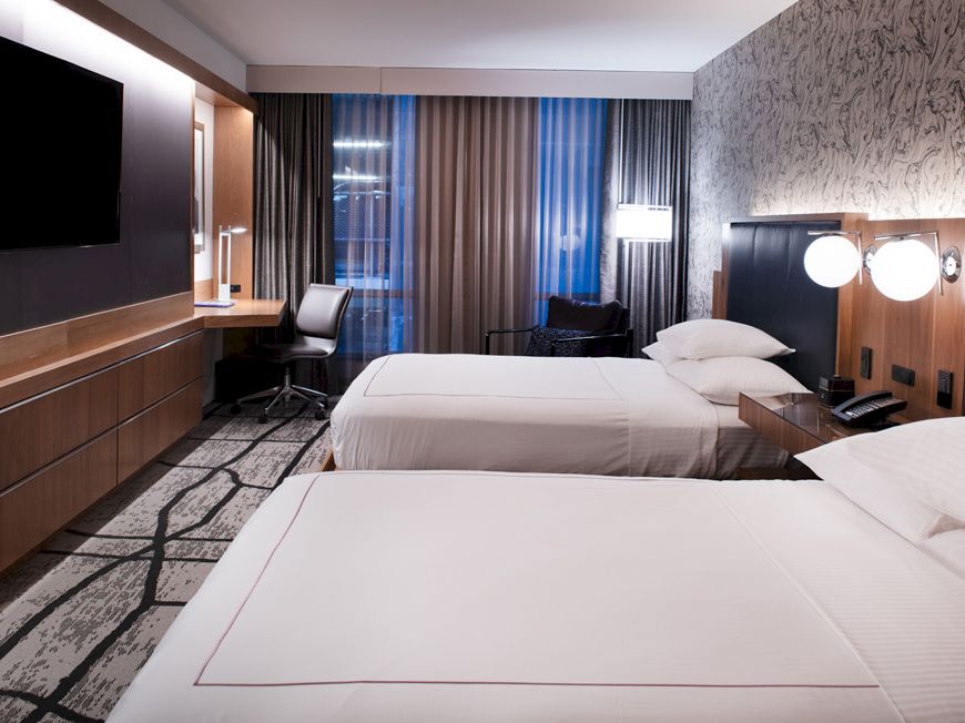 A modern hotel room features two twin beds, a wall-mounted TV, a desk with a chair, and floor-to-ceiling windows with curtains and city views.
