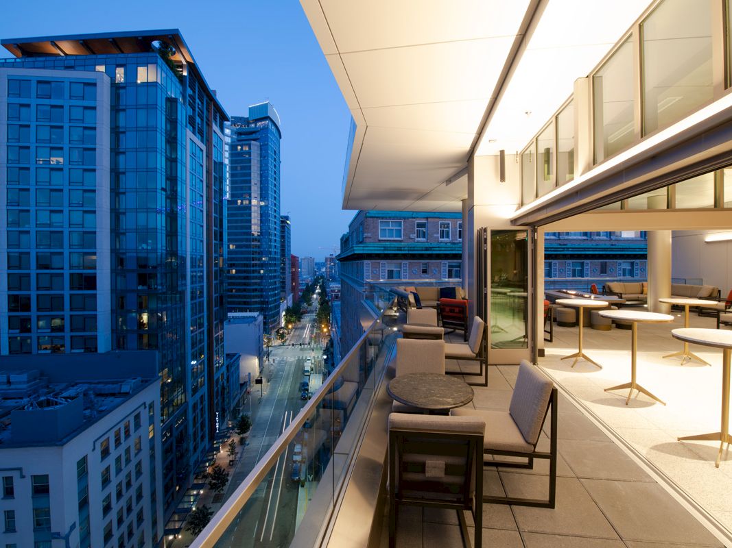 A modern cityscape at dusk viewed from a sleek balcony with outdoor seating, overlooking illuminated buildings and a bustling street below.